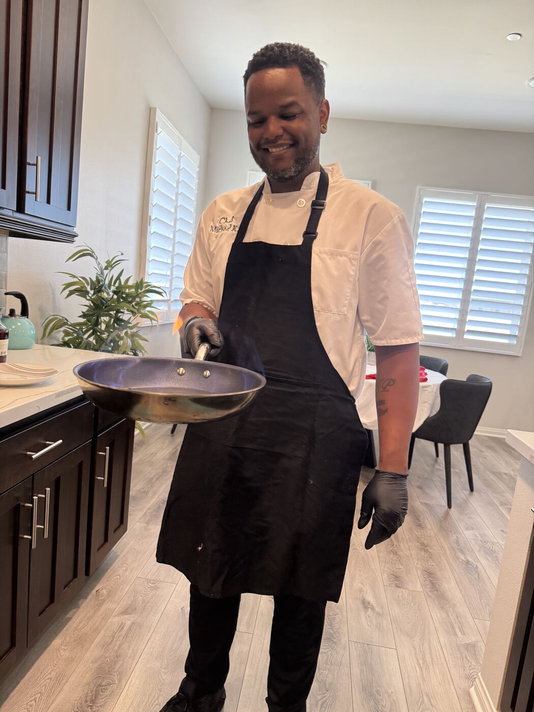 Chef Michael King smiling in his white chef coat, holding a flaming sauté pan in a client's home kitchen